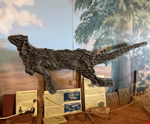 A dinosaur sculpture made of metal pieces, displayed against a mural of a prehistoric landscape.
