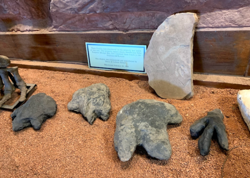 Display of ancient stone tools and artifacts on a sandy surface, with an informational sign in the background.