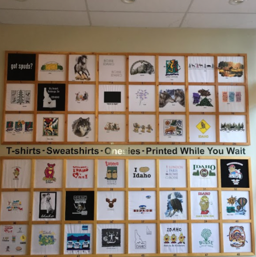 A wall display featuring various printed designs on t-shirts, sweatshirts, and onesies, showcasing Idaho-themed graphics.