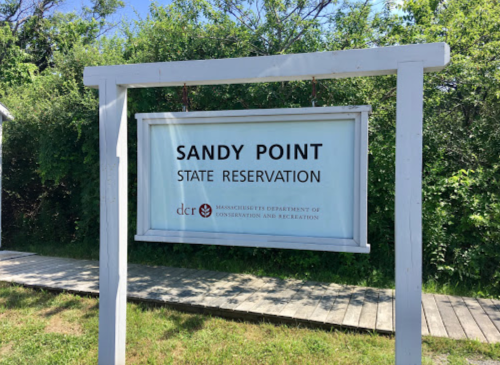 Sign for Sandy Point State Reservation, surrounded by greenery, with a wooden walkway in the foreground.
