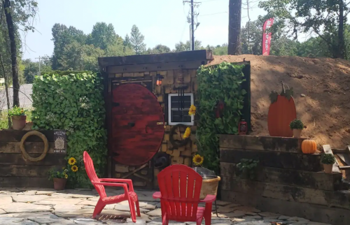 A whimsical outdoor scene featuring a red door surrounded by greenery, with red chairs and decorative pumpkins nearby.