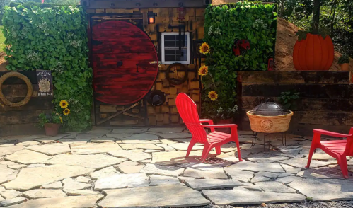 A whimsical outdoor space featuring a red door, stone patio, two red chairs, and vibrant greenery with sunflowers.