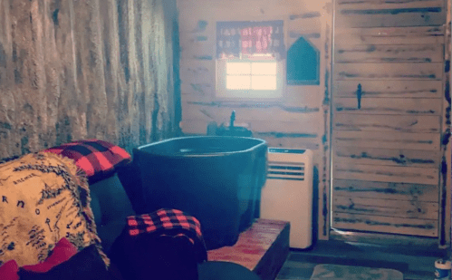 Cozy interior of a rustic cabin featuring a black tub, a couch with blankets, and a wooden door.