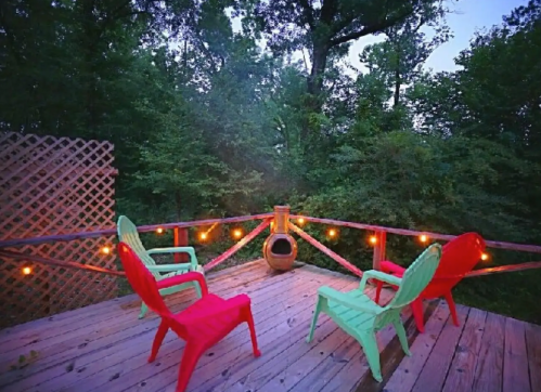 A cozy outdoor deck with red and green chairs, surrounded by trees and string lights, creating a warm atmosphere.