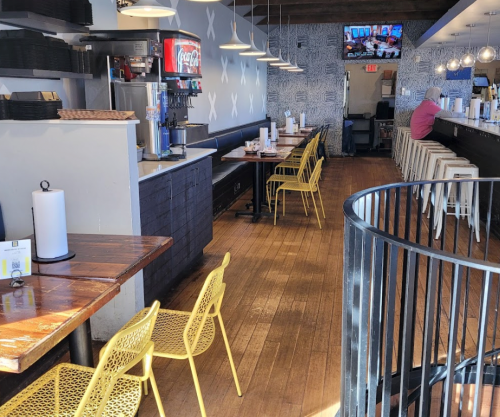 Interior of a restaurant featuring wooden tables, yellow chairs, a drink station, and a bar area with stools.