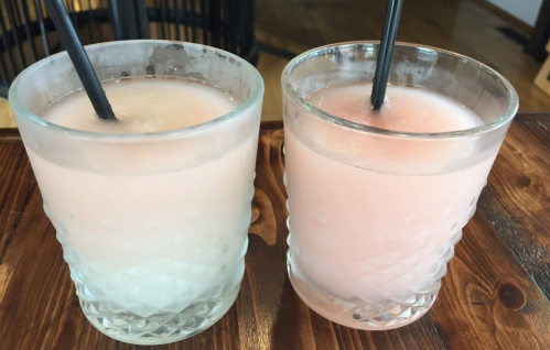 Two glasses of pink and light pink drinks with black straws, placed on a wooden table.