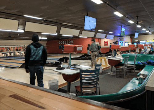 A bowling alley interior with people, lanes, and screens; one person stands while another walks away.