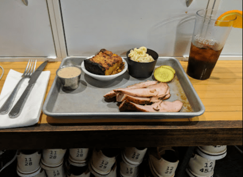 A metal tray with sliced meat, pickles, macaroni, cornbread, and a drink on a wooden table.