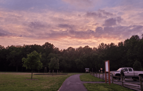 A serene park scene at sunset, featuring a path, trees, and a few parked cars under a colorful sky.