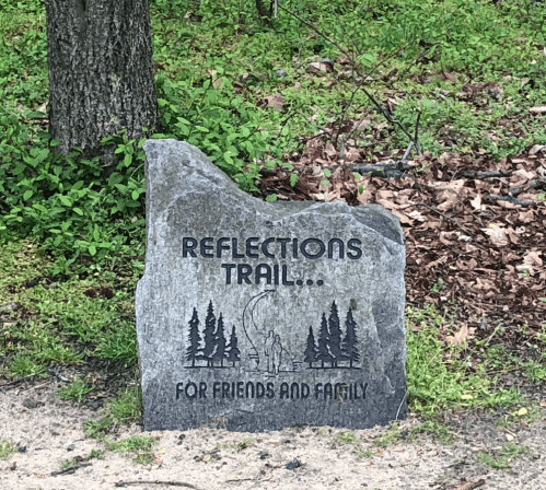 A stone marker for "Reflections Trail" surrounded by trees and leaves, dedicated to friends and family.