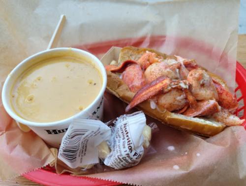 A bowl of clam chowder next to a lobster roll on a paper-lined tray, with a piece of candy on the side.