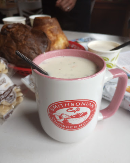 A mug of clam chowder with a pink rim, surrounded by bread and snacks on a table.