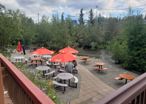 Outdoor seating area by a river, featuring tables with red umbrellas and surrounded by greenery.