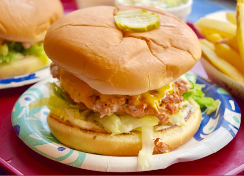 A close-up of a chicken sandwich with lettuce, cheese, and a pickle slice on a paper plate, with fries in the background.