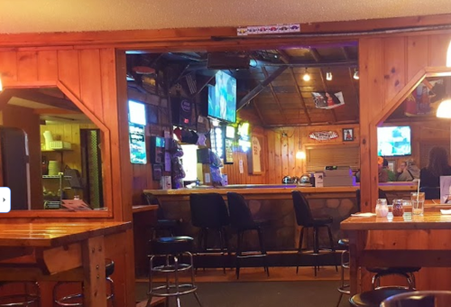 A rustic bar interior with wooden walls, stools, and multiple TVs displaying sports.