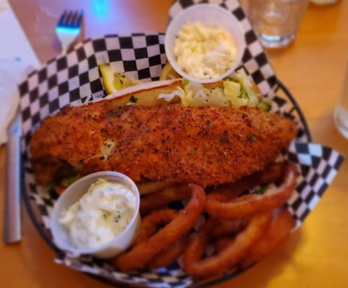 A plate with a large fried fish fillet, onion rings, and two small cups of dipping sauce on a checkered paper.