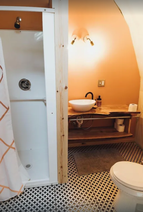 A cozy bathroom featuring a shower, wooden shelf, sink, and toilet with patterned floor tiles and warm orange walls.