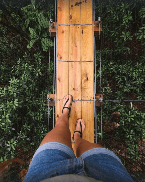 A person stands on a wooden bridge above lush greenery, wearing shorts and flip-flops.