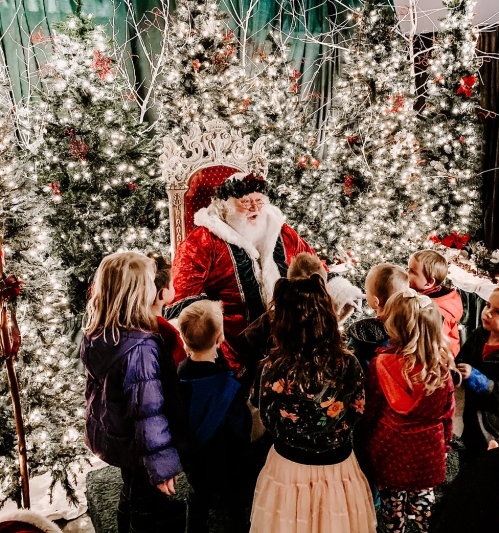 Santa Claus sits on a throne surrounded by children, with festive trees and lights in the background.