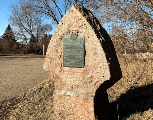 A large, weathered stone monument with a plaque, set against a backdrop of trees and a clear sky.