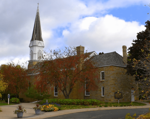 A stone church with a tall spire, surrounded by colorful autumn trees and a clear blue sky.