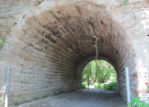 A stone archway leading into a lush, green park, with trees and a sign indicating park rules nearby.