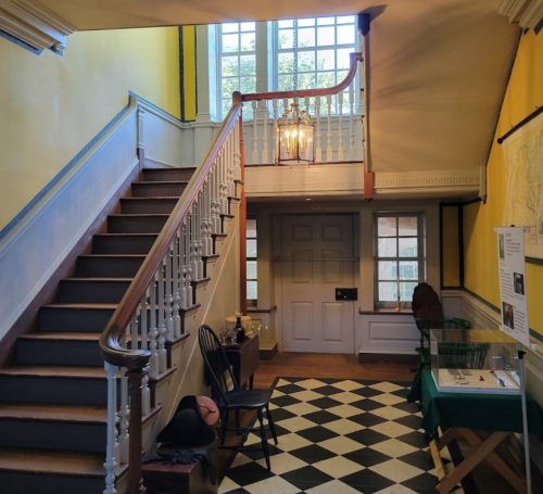 A bright foyer with yellow walls, a staircase, and a checkered floor, featuring a chandelier and wooden furniture.
