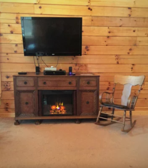 Cozy living room with a wooden TV stand, electric fireplace, and a rocking chair against a rustic wooden wall.