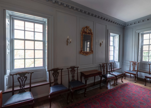A well-lit room with two windows, ornate mirror, and wooden chairs arranged along the walls.
