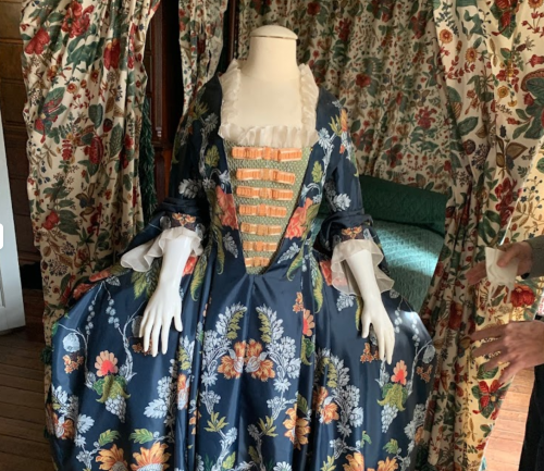 A vintage dress displayed on a mannequin, featuring floral patterns and intricate detailing, set against a patterned backdrop.