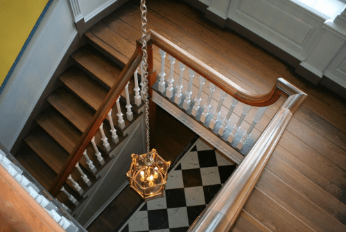 A view of a wooden staircase with a chandelier, featuring a patterned floor and white railings.