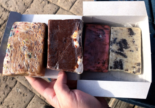 A hand holding a box containing four wrapped bars of different flavors or types of fudge.