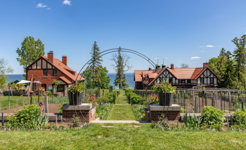 A scenic view of a garden with flower pots, leading to two charming houses by the lake under a clear blue sky.