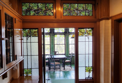 A view of a vintage kitchen with stained glass windows and a doorway leading to a sunlit dining area.