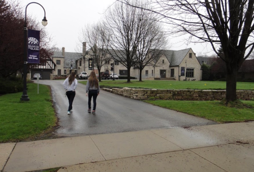 Two people walk down a paved path towards a large building, surrounded by trees and grass on a cloudy day.