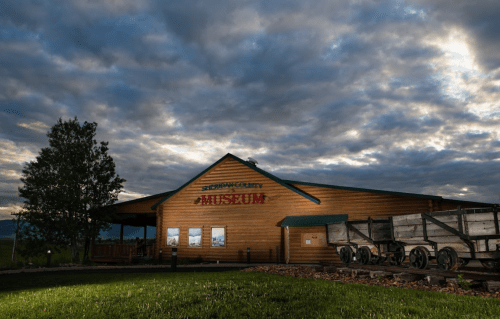 A wooden museum building with a sign, surrounded by clouds and a wagon display in front.