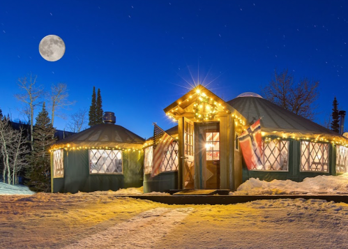 A cozy yurt decorated with lights and flags, surrounded by snow under a bright moonlit sky.