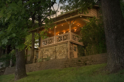 A cozy wooden house with a stone foundation, featuring a lit porch surrounded by trees and a grassy area.