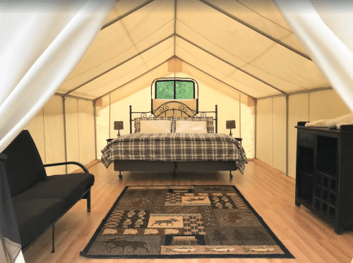 Cozy glamping tent interior with a double bed, two lamps, a sofa, and a decorative rug on wooden flooring.