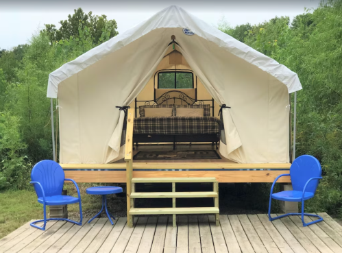A cozy glamping tent with a bed and two blue chairs on a wooden deck, surrounded by greenery.