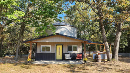 A charming round house with a yellow door, surrounded by trees and outdoor seating.
