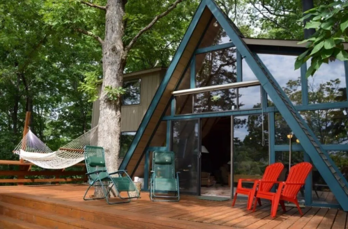 A modern A-frame cabin surrounded by trees, featuring a deck with red chairs and a hammock.