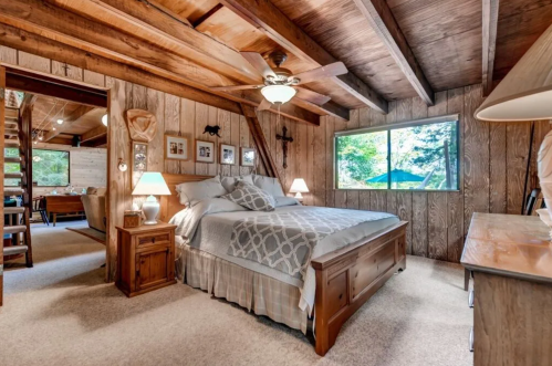 Cozy wooden bedroom with a large bed, ceiling fan, and windows overlooking a forested area. Warm, inviting decor.