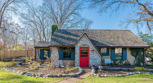 A charming stone cottage with a red door, surrounded by a landscaped garden and trees in the background.