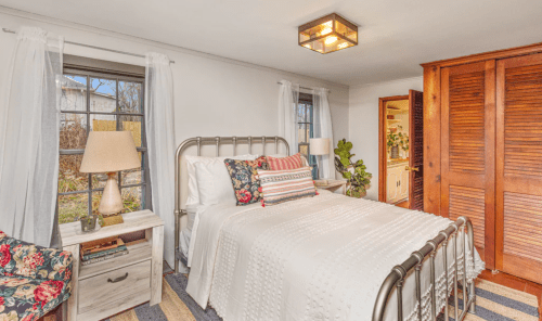 Cozy bedroom with a metal bed frame, floral accents, a nightstand, and natural light from windows.