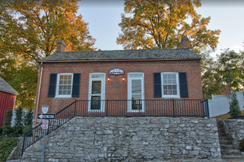 A brick house with black shutters, set on a stone foundation, surrounded by autumn trees.