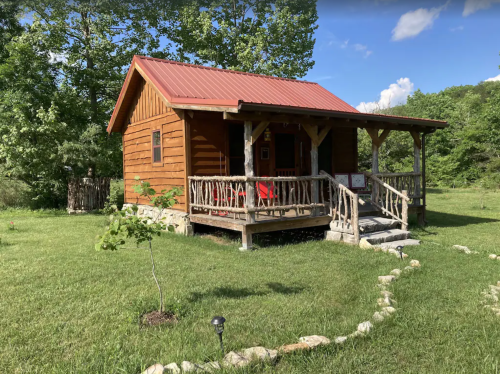 A cozy wooden cabin with a red metal roof, surrounded by greenery and a stone path leading to it.