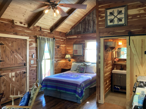 Cozy cabin interior featuring a bed, wooden walls, a ceiling fan, and a small bathroom area. Warm, rustic decor.