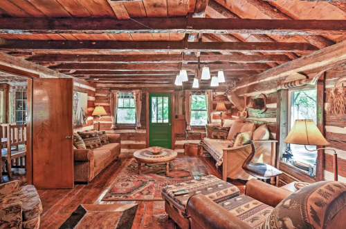 Cozy log cabin interior with wooden beams, comfortable seating, a round table, and large windows letting in natural light.