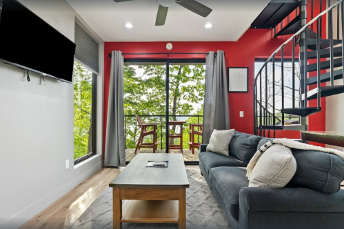Cozy living room with a gray sofa, wooden coffee table, and large windows overlooking greenery. Spiral staircase in the background.